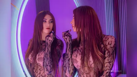 AndreaKalo with long brown hair wears a black snakeskin mesh bodysuit while looking into a circular purple-lit mirror.