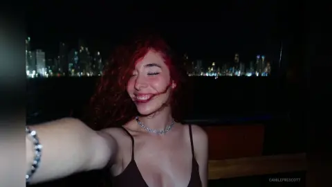 Smiling woman with curly red hair wearing a dark camisole on a rooftop balcony overlooking a city skyline at night.