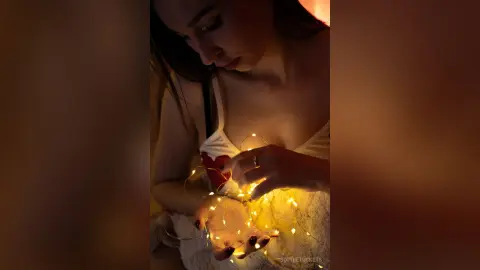 Young woman in a white lace dress holding glowing fairy lights in a warm, moody indoor setting.