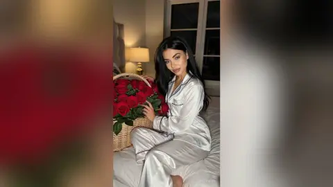 Beautiful woman in white silk pajamas sitting on a bed holding a large basket of red roses in a cozy bedroom.