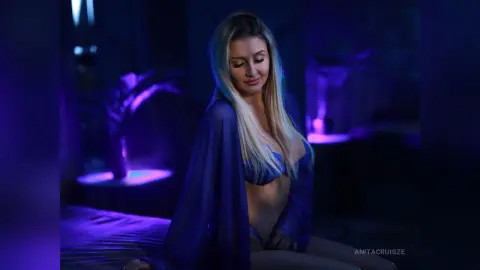 Anita wears a blue lace bra and matching sheer robe while sitting in a dark, purple-lit room.
