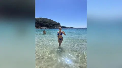 Young woman in a blue bikini standing in clear turquoise ocean water under a bright sunny sky.