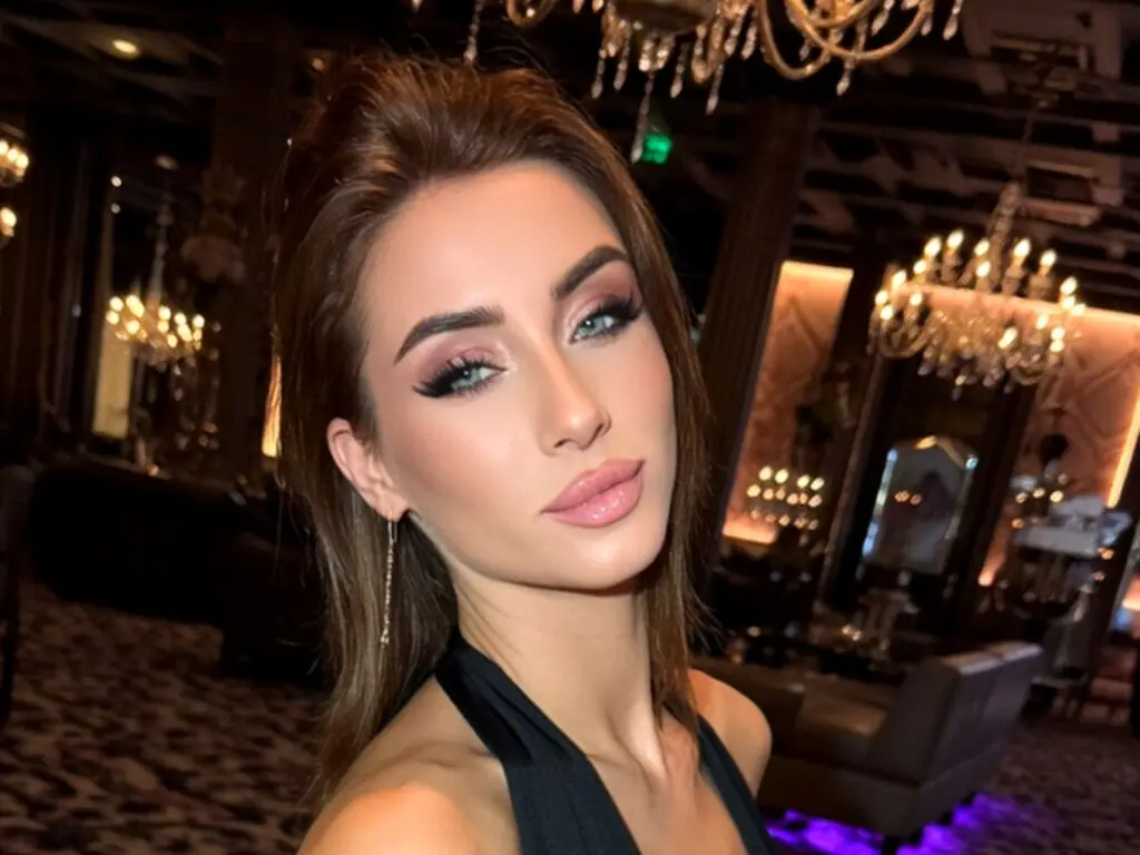 Glamorous woman with elegant makeup and dark dress posing in a luxury restaurant with chandeliers.