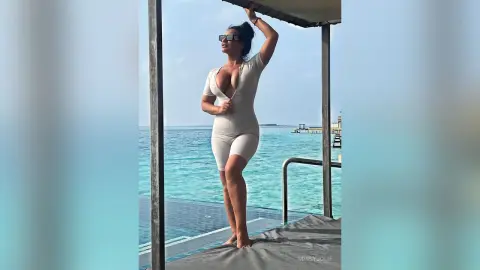 MissyJolie stands on a deck overlooking the ocean wearing a tight white bodysuit and sunglasses.