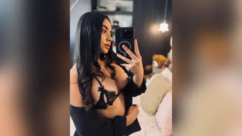 Young woman with long dark hair wearing a black lace bra and blazer in a cozy bedroom setting.