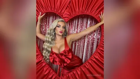 Glamorous blonde woman in a red satin dress posing inside a large red heart backdrop for a romantic photoshoot.