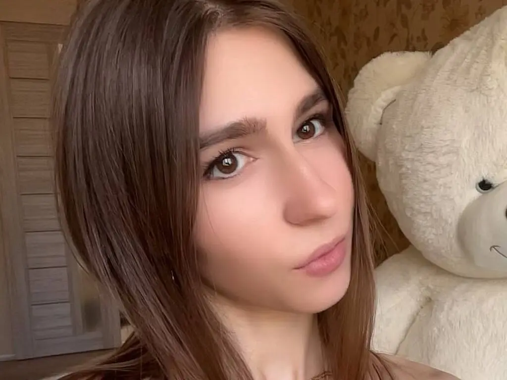 Young woman with long brown hair posing indoors next to a large white teddy bear in a cozy setting.