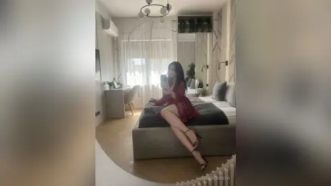 Young woman in a red silk robe sitting on a bed in a bright, modern bedroom taking a mirror selfie.