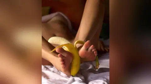 VeraSurikova lies on a white bed sheet, holding a partially peeled banana near her bare feet and legs.