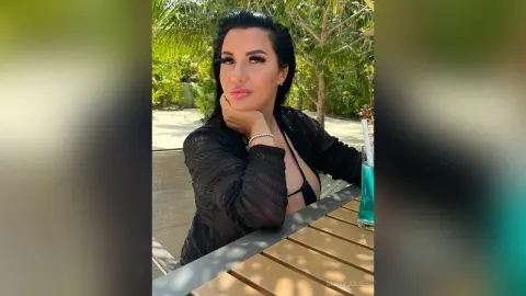 MissyJolie poses outdoors in a black sheer top, resting her chin on her hand near a blue cocktail.