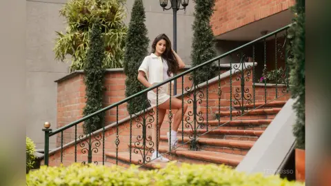 TefyRusso poses on outdoor brick stairs wearing a white short-sleeved top and white shorts, standing next to a green metal railing and tall green bushes.