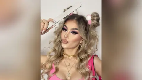 Glamorous blonde woman with wavy hair and gold jewelry posing in a pink top against a white background.