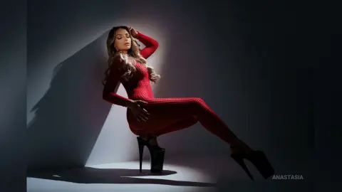 Anastasia poses in a tight red long-sleeved bodysuit with her leg extended against a plain wall.