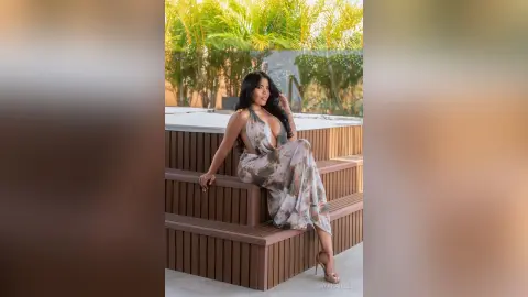 DayanaHille sits on a wooden deck wearing a long, patterned sundress with a deep plunging neckline.
