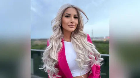 Blonde woman in a pink blazer and white crop top posing outdoors against a soft blue sky background.