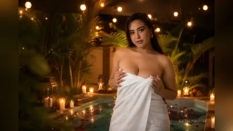 NatalyBloom poses wrapped in a white towel by a lit pool at night.