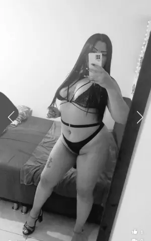 Black and white mirror selfie of a woman in black bikini posing in a bedroom setting.