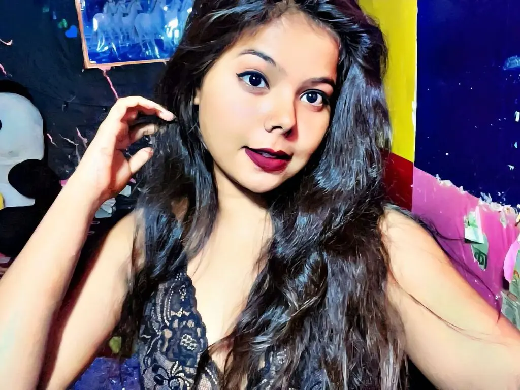 Young woman with long wavy hair wearing a black lace top in a colorful indoor setting.