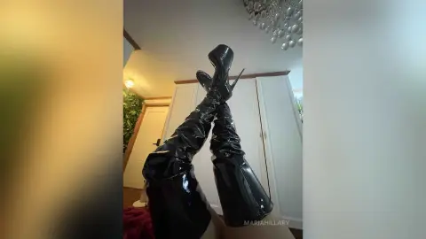 Woman wearing shiny black patent leather thigh-high boots in a bright modern bedroom setting.