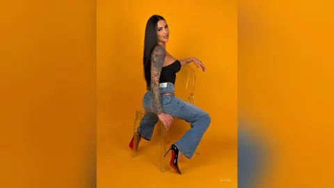 Tattooed woman in black crop top and blue jeans sitting on a clear chair against a vibrant yellow background.