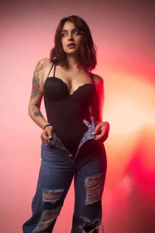 Young woman with tattoos in a black bodysuit and ripped jeans posing against a warm pink studio background.