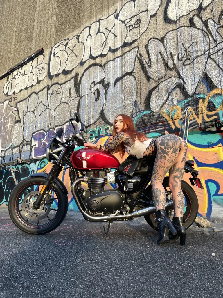 Lunaxluck poses with her long red hair and arm tattoos, wearing a black leopard print top against a warm marble background.
