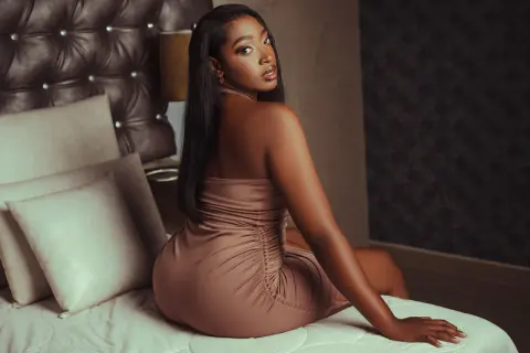 Elegant woman in a mocha satin dress sitting on a bed in a moody, luxurious bedroom setting.