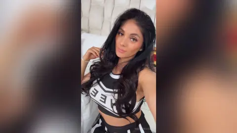 Young woman with long dark hair wearing a black and white crop top in a bright bedroom setting.