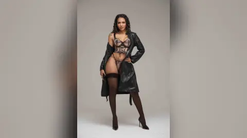 Confident woman in black leather jacket and lace lingerie posing in a minimalist studio setting.