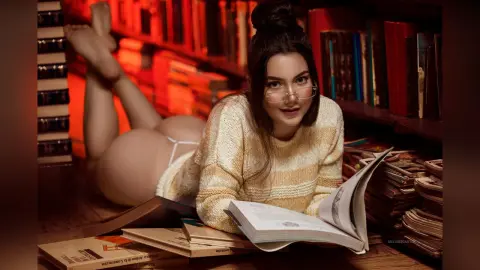 Young woman in a cozy cream sweater reading a book on a library floor with warm, moody lighting.