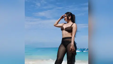 Smiling woman in a brown bikini and black sheer beach cover-up posing on a sunny tropical beach.