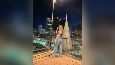 Stylish woman in a black crop top and baggy jeans posing on a city rooftop at night with skyscrapers in the background.