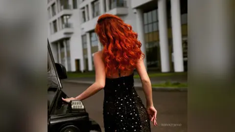 RubyAttwood stands with her back to the camera, wearing a black sequined dress, with her long red wavy hair falling down her back next to a black car.