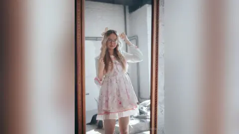 Young woman in a floral print dress posing in a mirror within a bright, minimalist room.