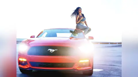 DrellaWolf — Stylish woman in a blue crop top and jeans posing on a red Ford Mustang under bright sunlight.