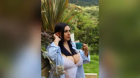 Young woman in a white lace bodysuit and blue shirt holding a teal mug on a lush green balcony.