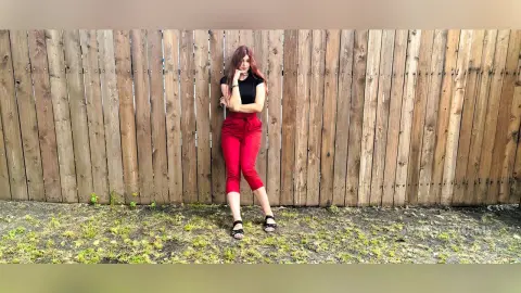 Stylish woman in a black top and red trousers leaning against a rustic wooden fence in soft daylight.