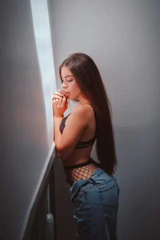 Young woman in a black bralette and denim jeans posing thoughtfully by a window in soft natural light.