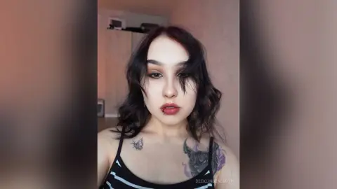 Young woman with wavy dark hair and tattoos wearing a black striped tank top in a soft indoor setting.