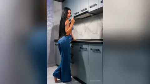 Long-haired woman in blue denim outfit standing in a modern kitchen with grey cabinets.