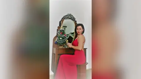 Elegant woman in a long red evening gown sitting by a vintage vanity mirror in a bright room.