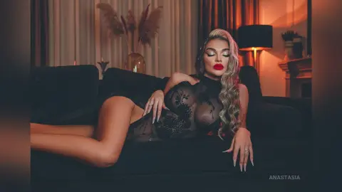 Anastasia reclines on a black sofa wearing a black lace bodysuit that shows her breasts and legs.