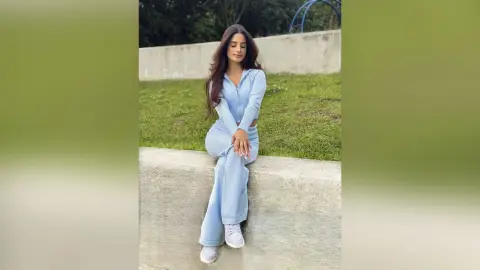 TefyRusso sits on a concrete ledge wearing a light blue long-sleeve crop top and matching flared pants, showing off her long brown hair.