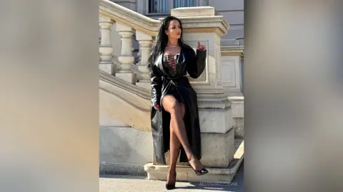 Elegant woman in a long black leather coat and heels posing by a classic stone staircase in sunlight.