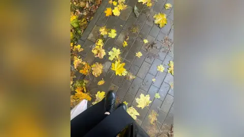 Person in black trousers walking on a wet brick path covered in bright yellow autumn maple leaves.