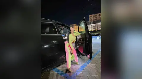 NicoleBella stands by an open car door wearing a lime green bodysuit and pink pants, posing against a dark night background.