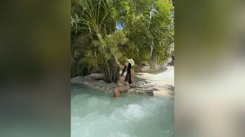 Aleja sits on the edge of a bright blue pool surrounded by lush green palm trees, wearing a light-colored bikini.