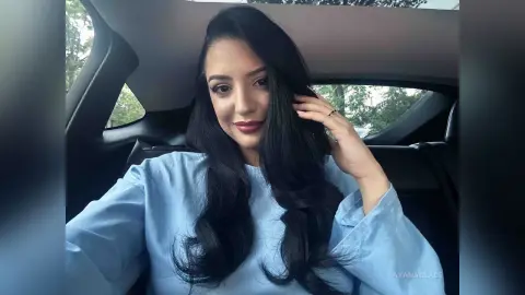 Smiling woman with long black hair wearing a light blue shirt sitting inside a car.