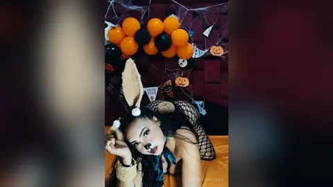 Woman in bunny ears and fishnet outfit posing playfully near orange and black Halloween balloons.