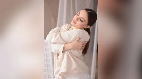 Serene young woman in a cream linen shirt embracing herself in a soft, dreamy indoor setting.